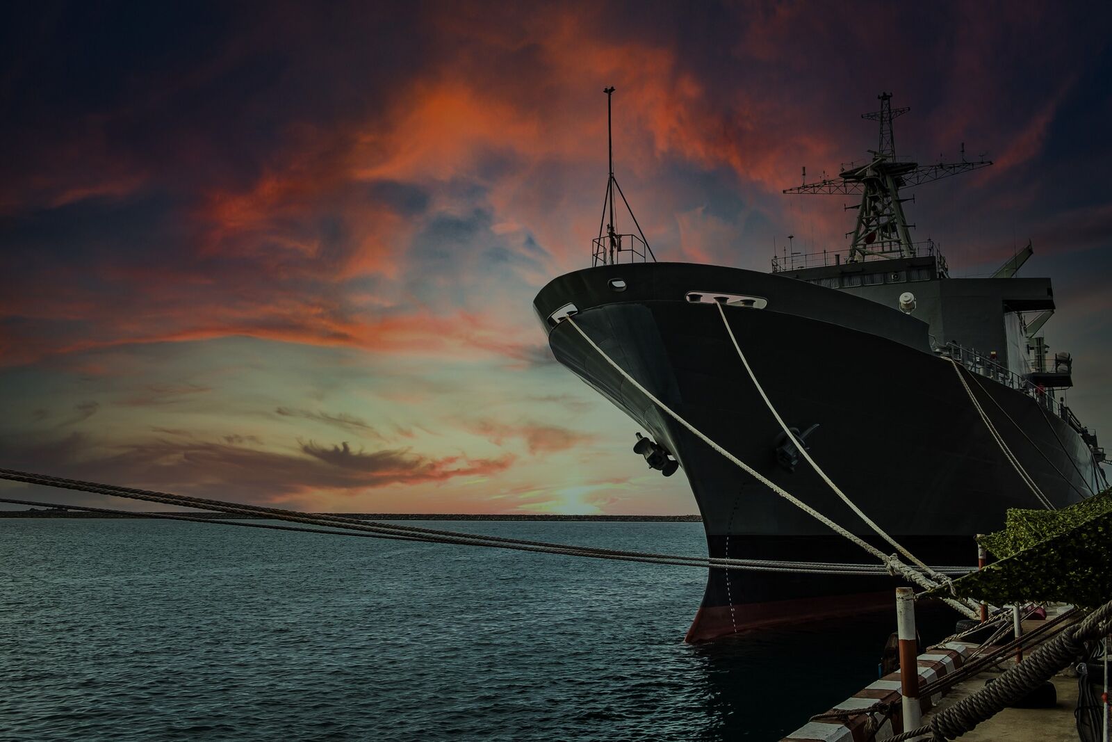 Naval vessel moored at a pier at sunset, with its bow and mooring lines in the foreground